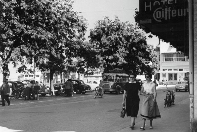 Elegante Passantinnen und historisches Postauto in einer lebendigen Strassenszene